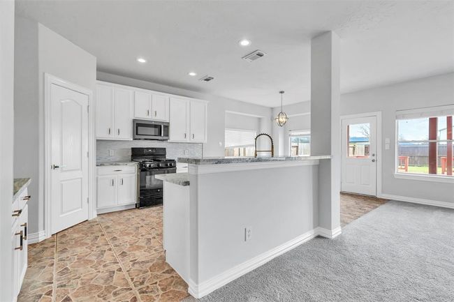 This photo showcases a bright, modern kitchen with white cabinetry and stainless steel appliances. It features a breakfast bar with granite countertops and an adjacent dining area with large windows offering natural light. The flooring transitions from tile in the kitchen to carpet in the living space, leading to a door that opens to the backyard. | Image 12
