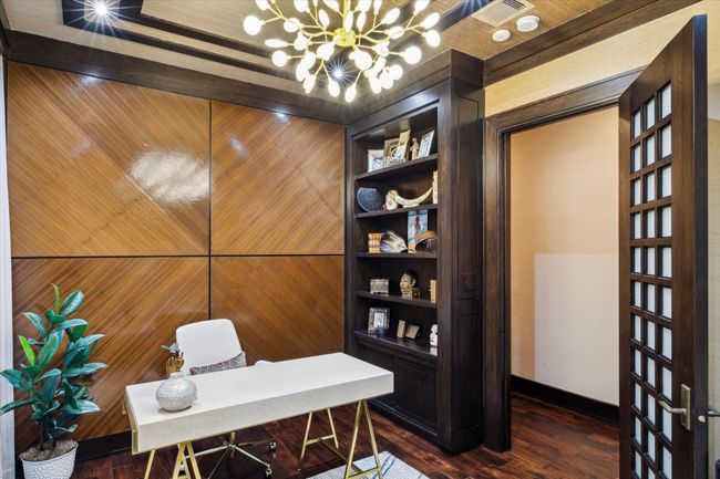 This additional angle of the secondary office showcases the room’s deeply grained wood-paneled feature wall and built-in display shelving, perfect for showcasing books, art, or personal mementos. The modern sculptural chandelier adds a modern touch of whimsy, while rich trim work and the 8-foot custom wood and milk glass door ground the space in warmth and sophistication. Whether used as a private study or reading room, this Zen-inspired space is as functional as it is visually impressive. | Image 24